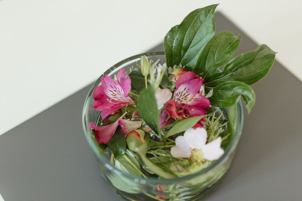 Sklenená nádoba s farebnými kvetmi a zelenými listami na stole.