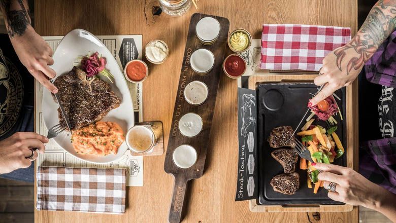 Dve osoby jedia steaky s prílohami pri drevenom stole.