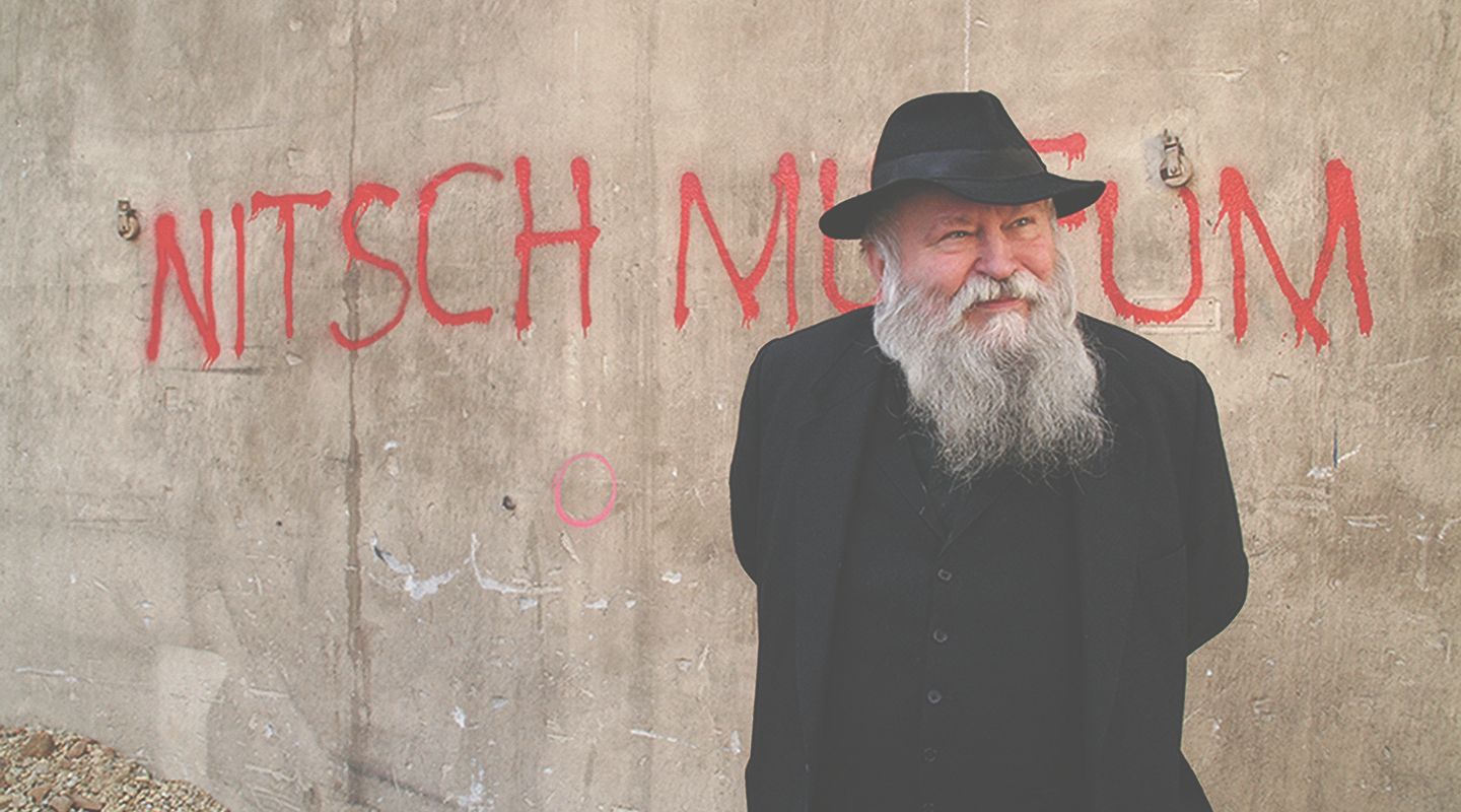 Muž s bradou a klobúkom stojí pred stenou s nápisom "Nitsch Museum".