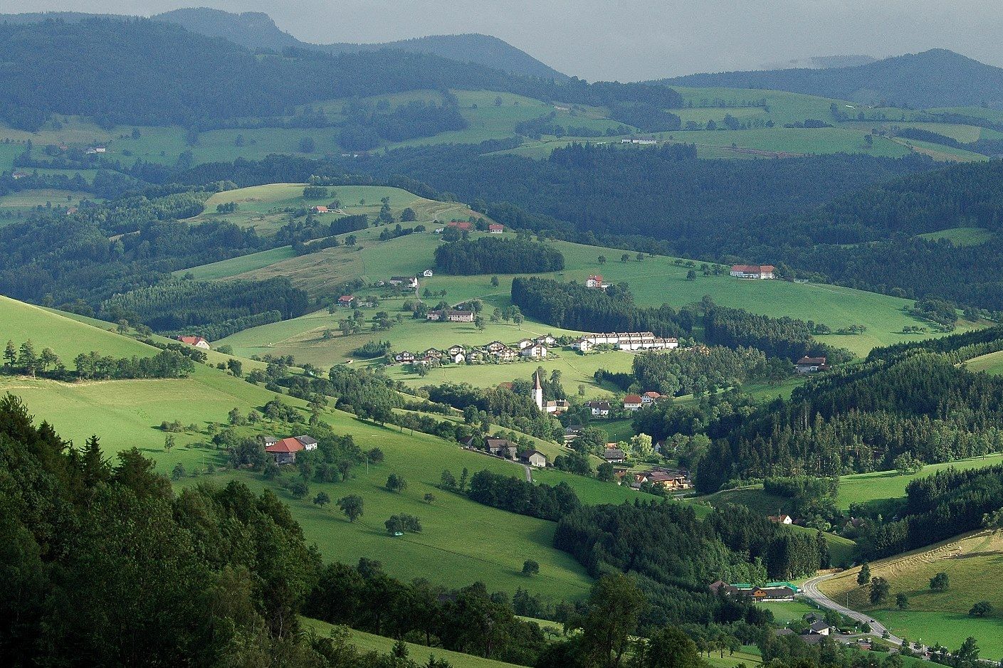 Panoramatický pohľad na zelenú kopcovitú krajinu s roztrúsenými domami a lesmi.