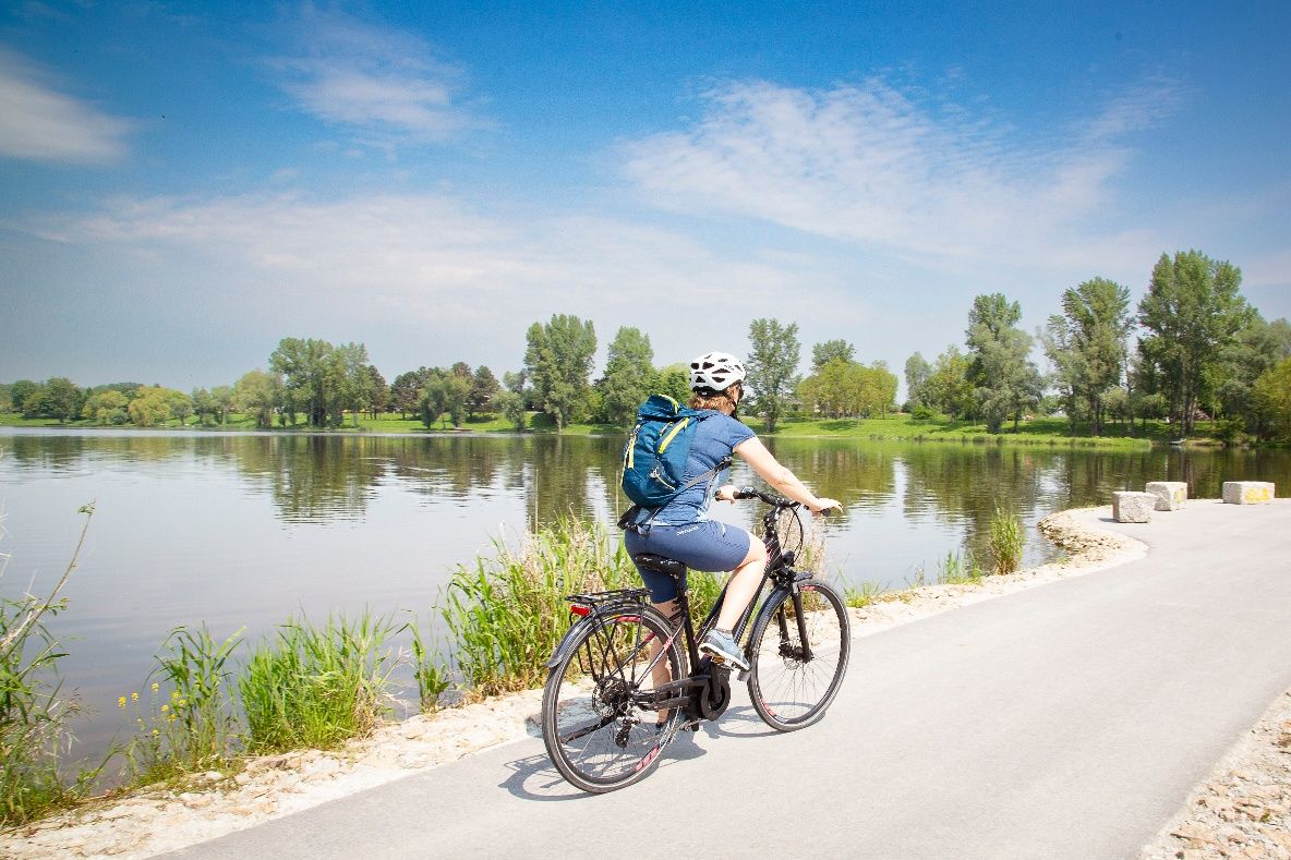 Osoba jazdí na bicykli po ceste pozdĺž rieky obklopenej zelenou krajinou a stromami.