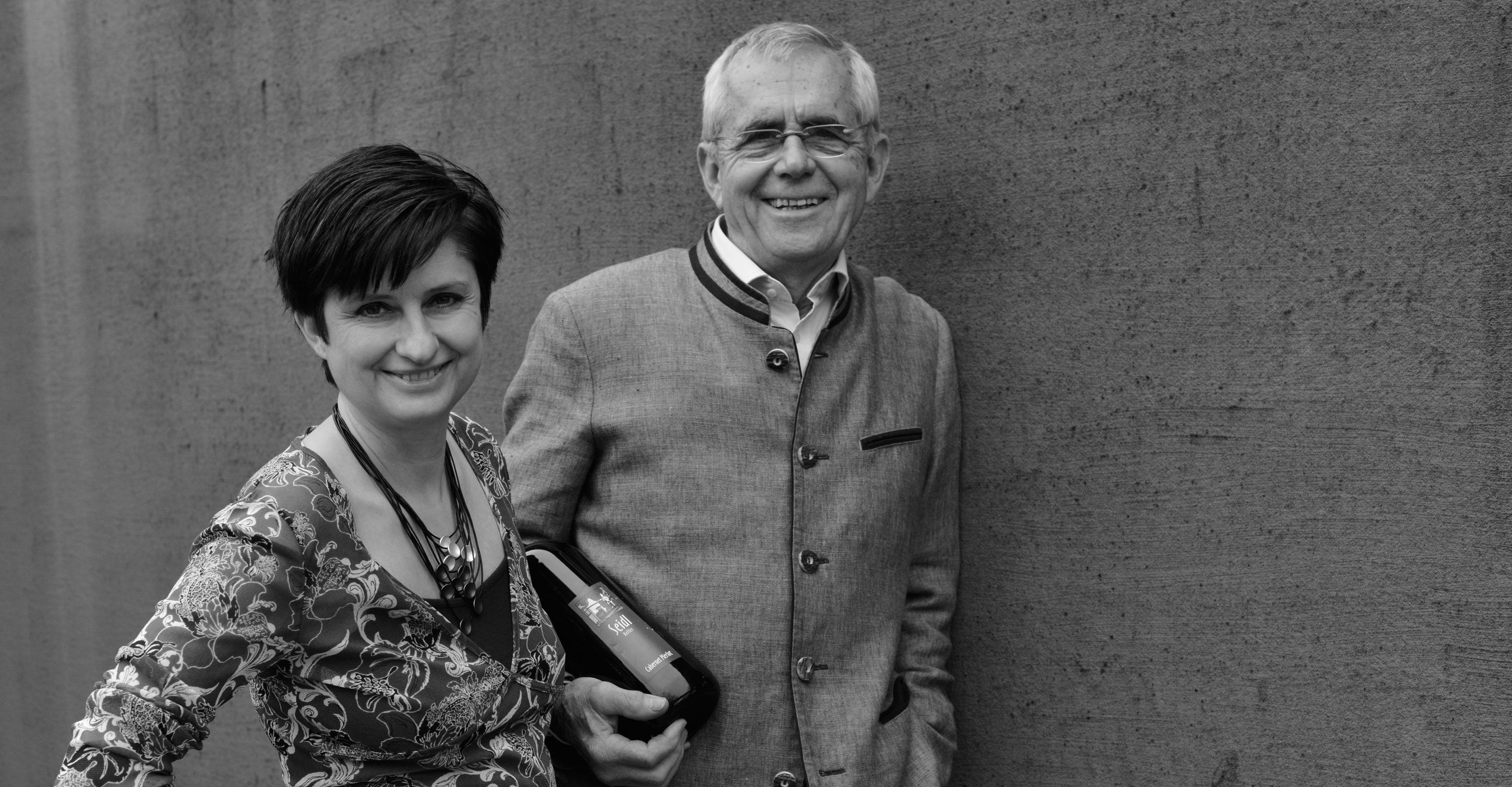 Muž a žena stoja s úsmevom pred stenou. Muž drží fľašu vína.