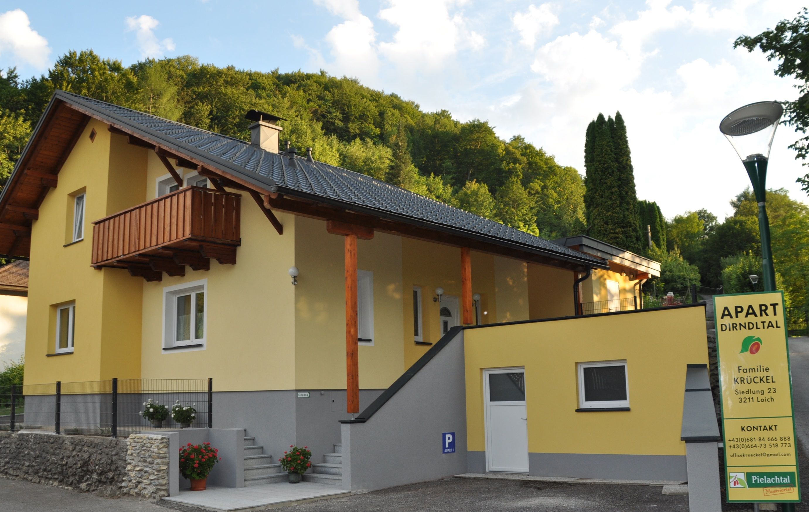Žltý dom s balkónom a nápisom "Apart Dirndltal" pred zalesneným kopcom.