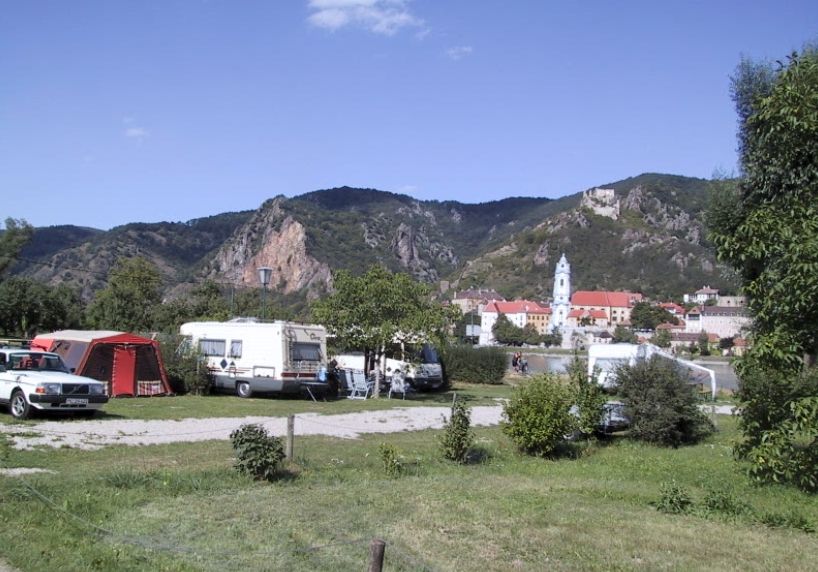 Kemp s karavanmi a stanmi pred horskou krajinou a kostolom v pozadí.