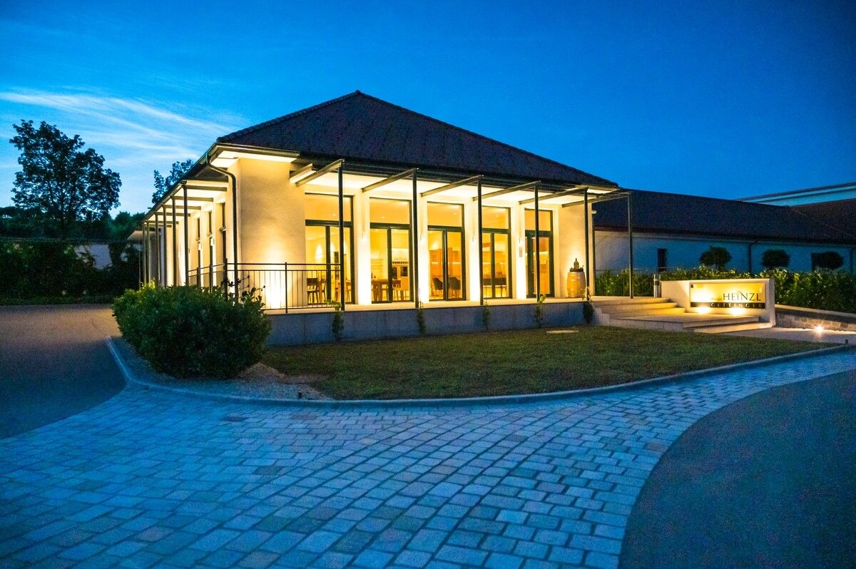 Moderná budova so sklenenými priečeliami, osvetlená za súmraku, obklopená dláždeným chodníkom a zelenými plochami.