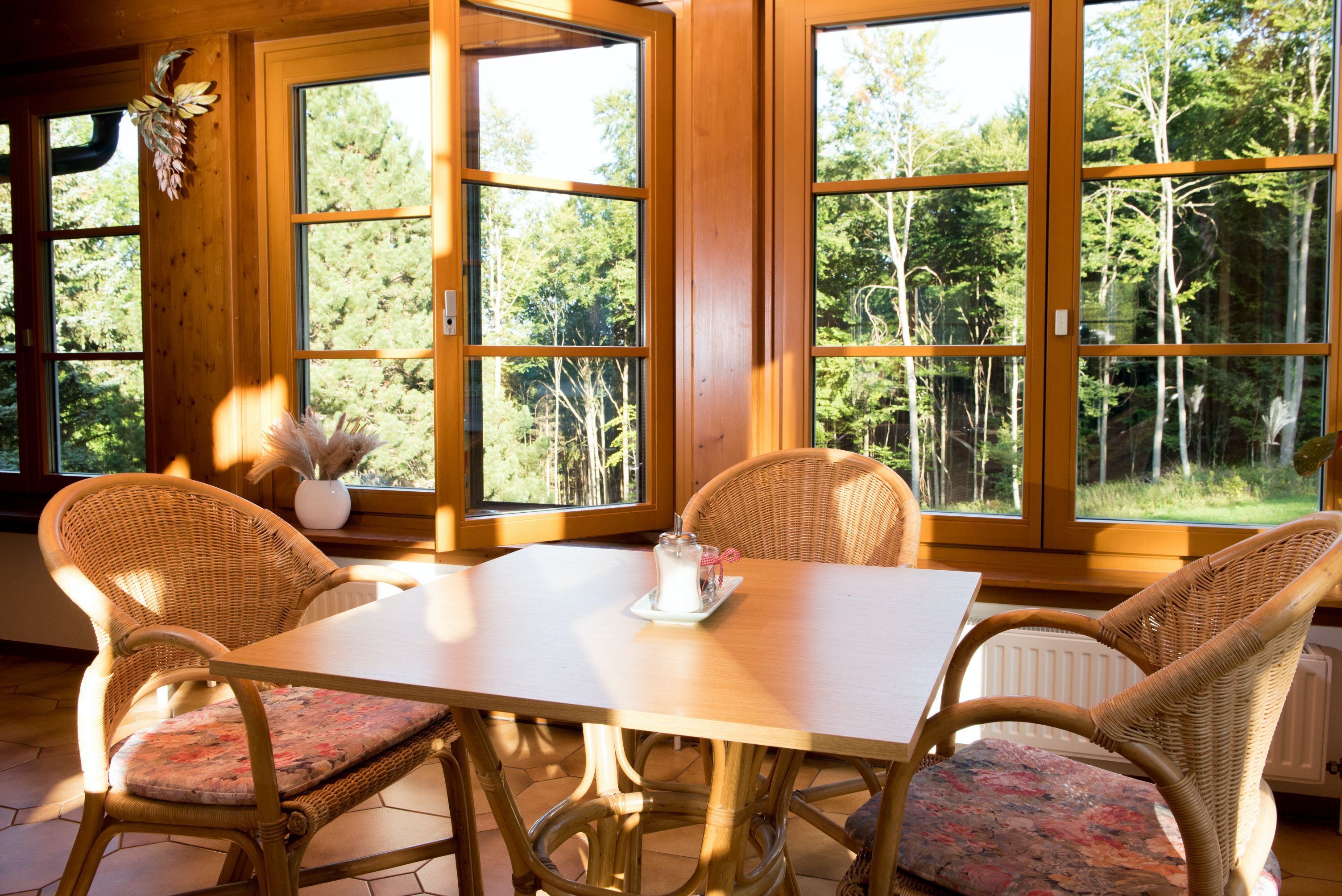 Stôl so štyrmi ratanovými stoličkami v slnečnej miestnosti s veľkými oknami a výhľadom na les.