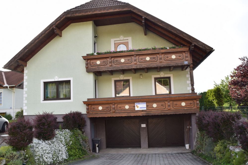 Dvojposchodový dom so zelenou fasádou a drevenými balkónmi, obklopený udržiavanými záhradami.