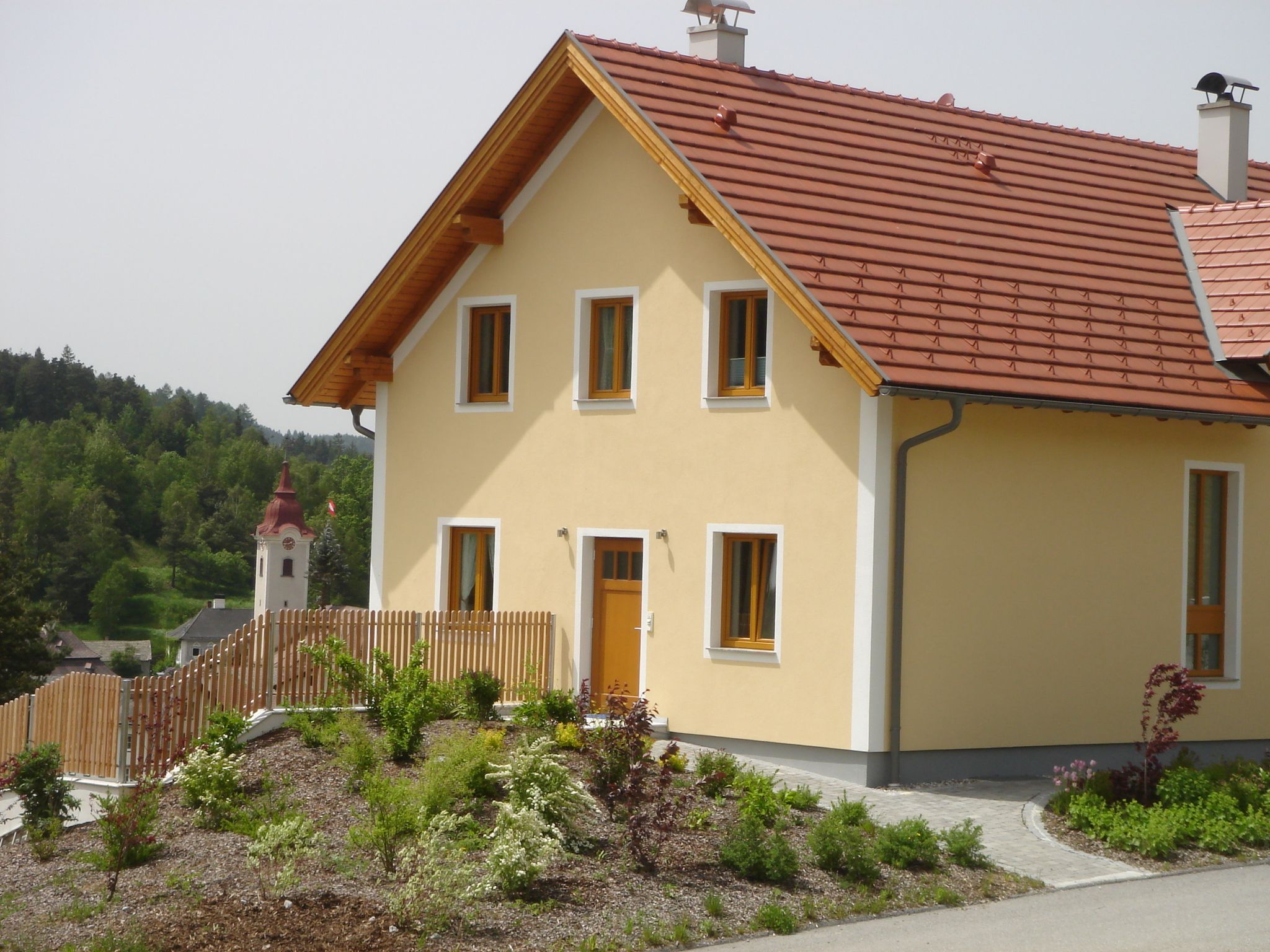 Moderný žltý dom s červenou strechou a záhradou, v pozadí kostol a zelená krajina.