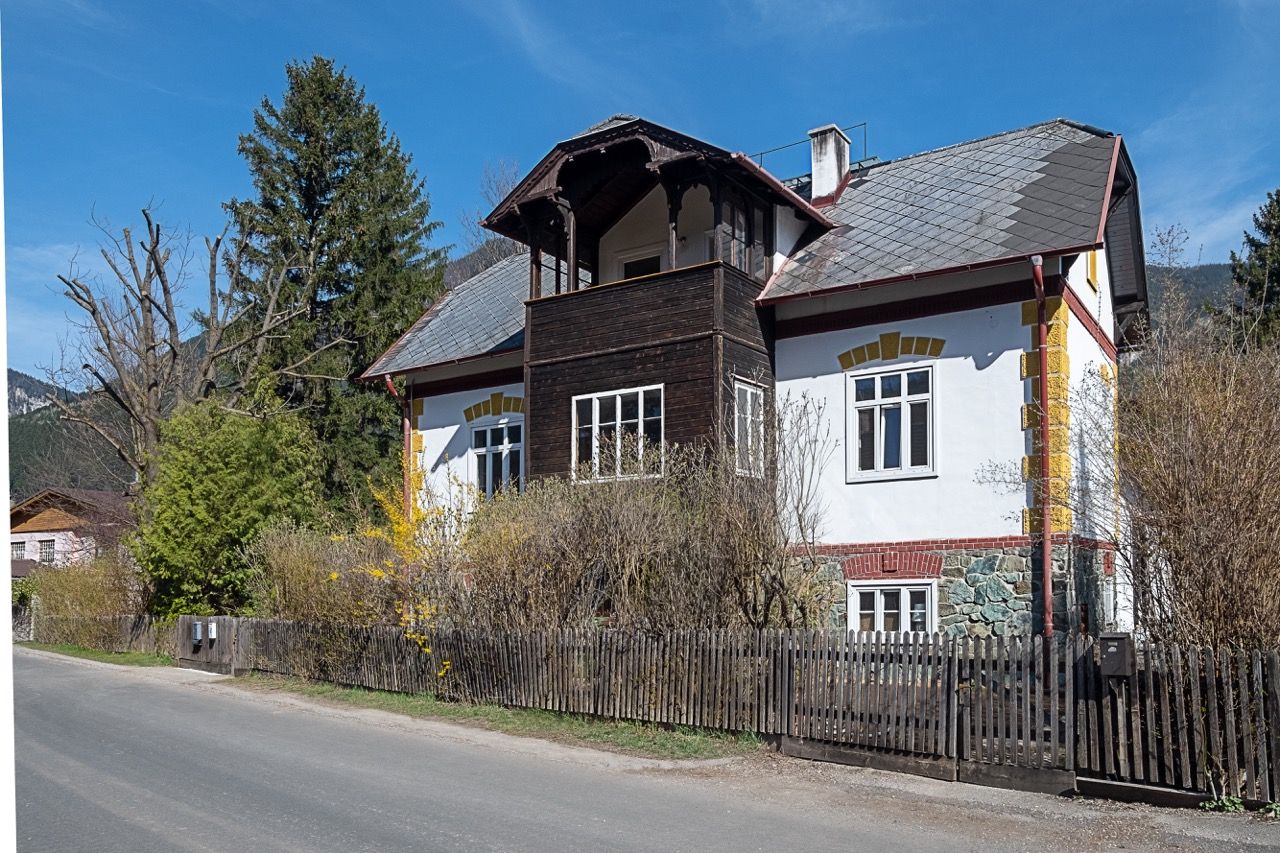 Tradičný vidiecky dom s dreveným balkónom a bielou fasádou, obklopený stromami a kríkmi, pod jasnou modrou oblohou.