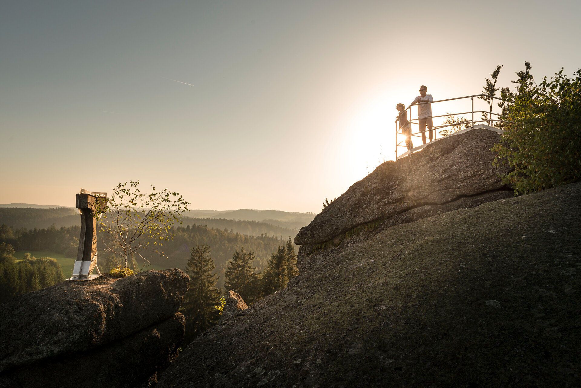 Zlaté večerné slnko zalialo majestátne skaly teplým svetlom, zatiaľ čo dvaja turisti si vychutnávajú úchvatný výhľad. Toto miesto obklopené nedotknutou prírodou a jemným šumením stromov vás pozýva na oddych a spoznávanie krásy hôr.