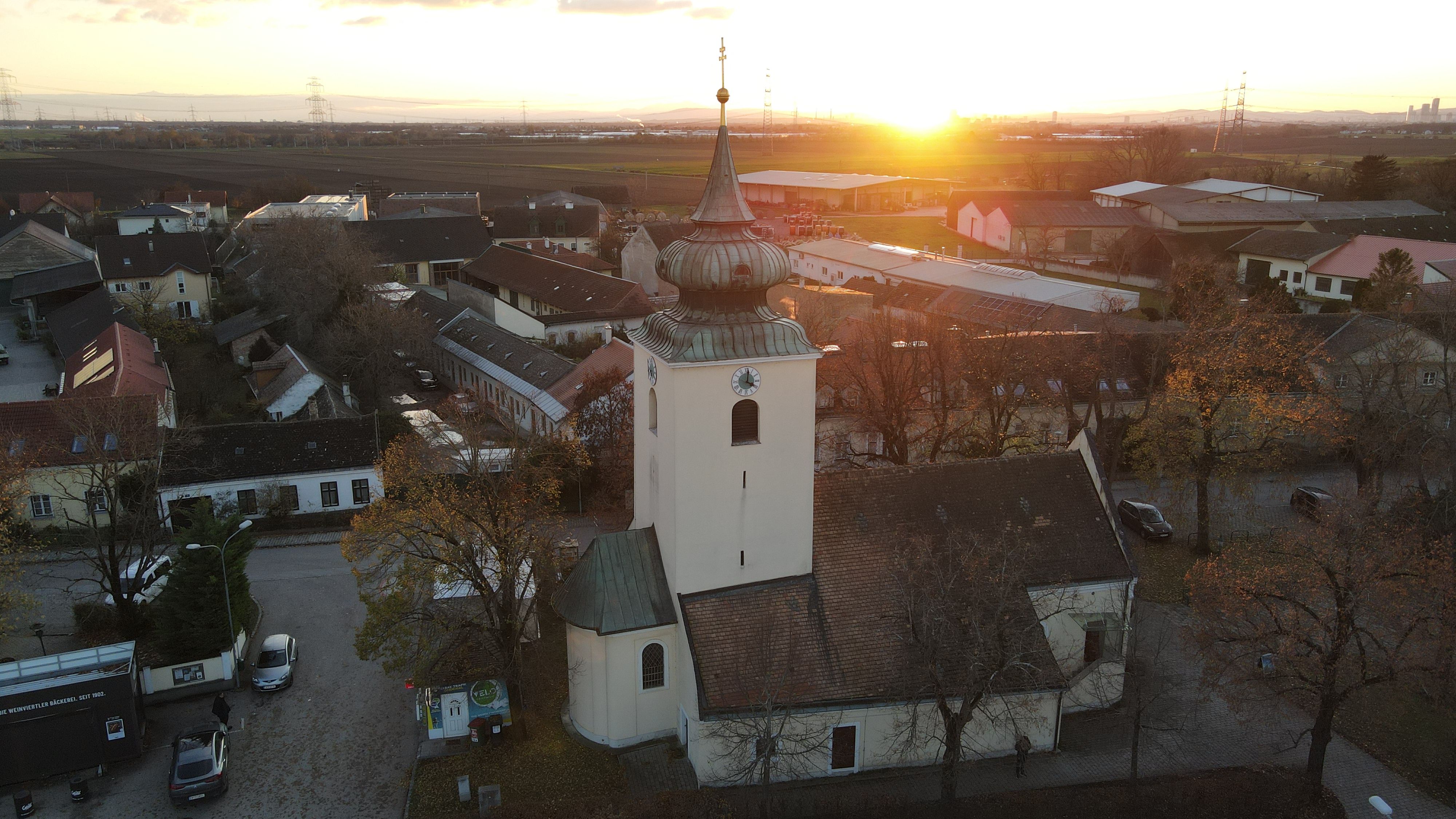 Kostol v Raasdorfe s vežou a cibulovitou strechou pri západe slnka, obklopený domami a poľami.