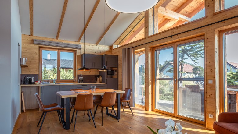 Pohľad do interi&eacute;ru modernej chaty s dreven&yacute;mi tr&aacute;mami, jed&aacute;lensk&yacute;m stolom a veľk&yacute;mi oknami.