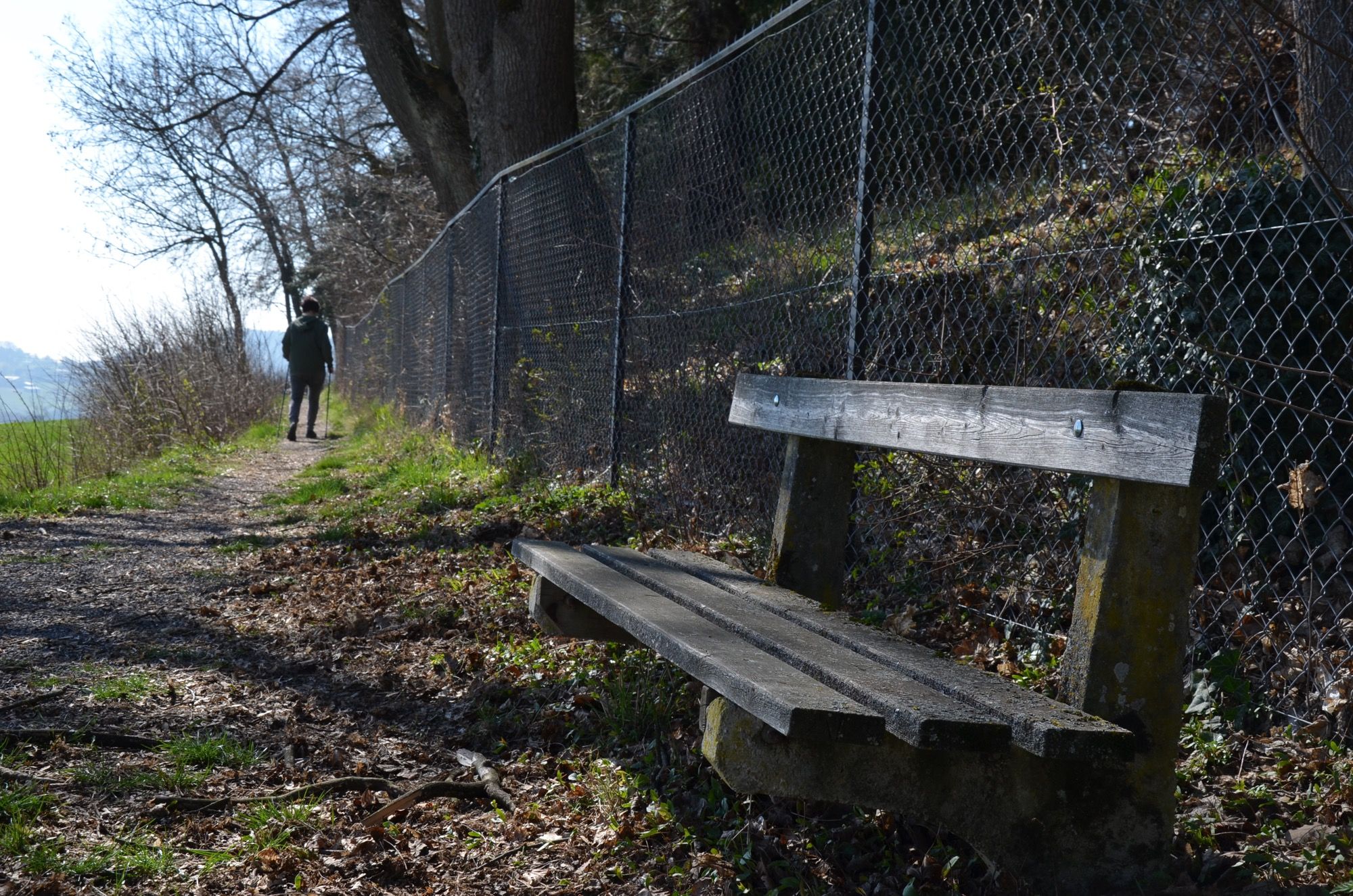 Chodník s drevenou lavičkou a plotom z drôteného pletiva. V diaľke kráča človek.
