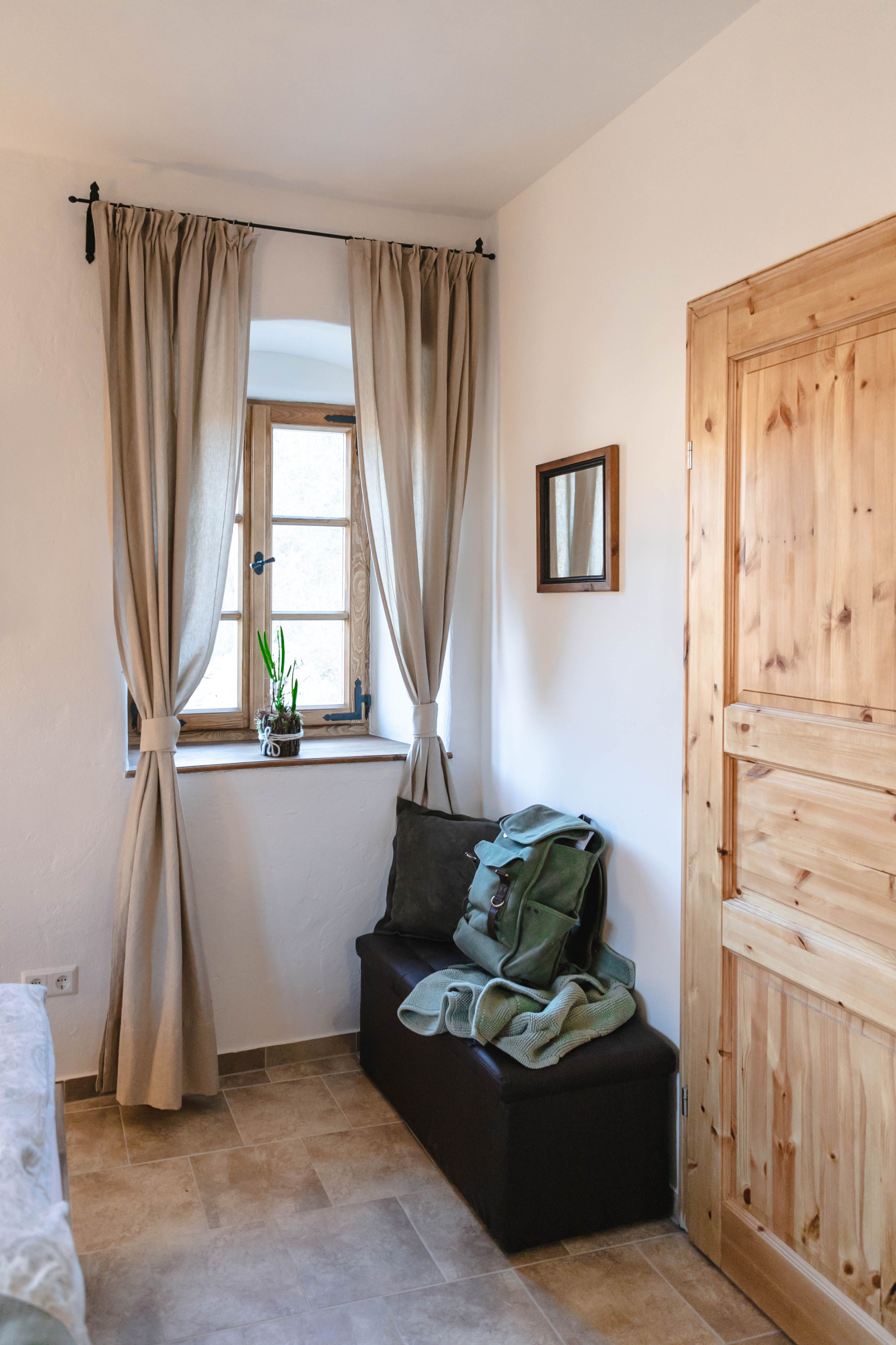 Malá predsieň s oknom, závesmi, drevenými dverami a lavičkou s batohom a dekou.