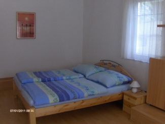 Jednoducho zariadená spálňa s manželskou posteľou, nočným stolíkom a oknom so závesmi.