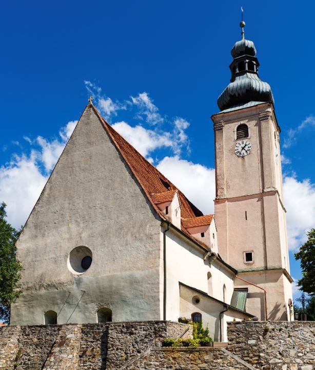 Farský kostol Wiesmath s vežou, kamenným cintorínskym múrom a hodinami na pozadí modrej oblohy.