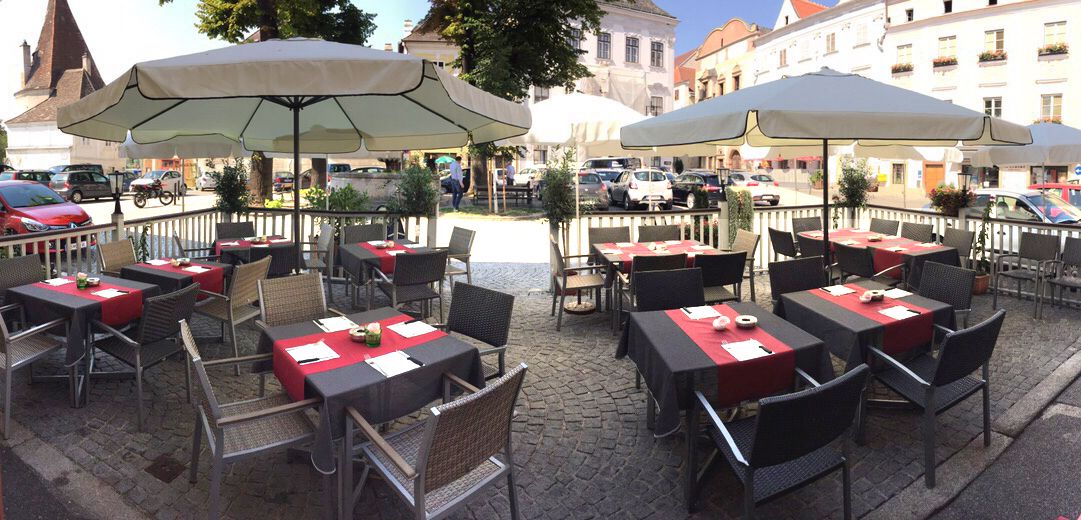 Vonkajšia terasa kaviarne so stolmi a slnečníkmi na vydláždenom námestí.