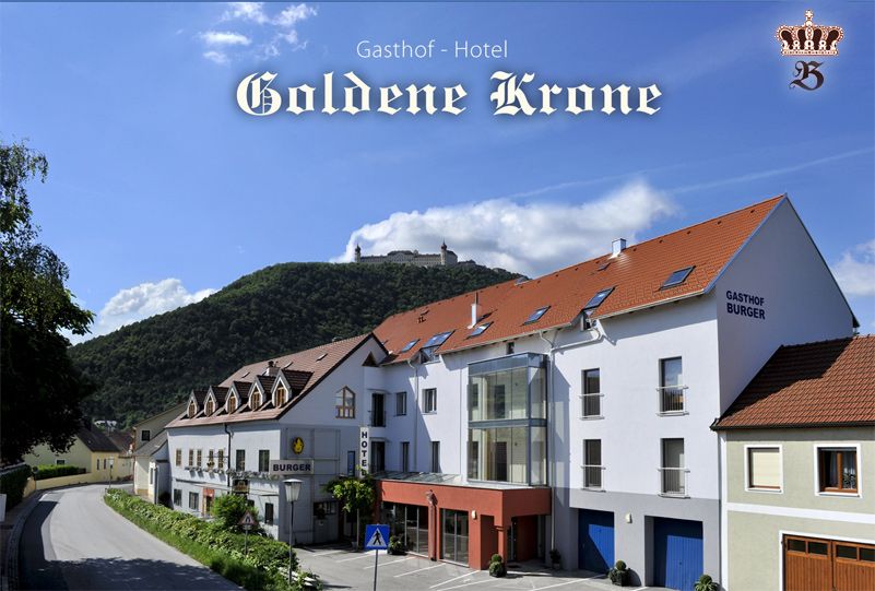 Gasthof-Hotel Goldene Krone pred zalesneným kopcom s hradom v pozadí.
