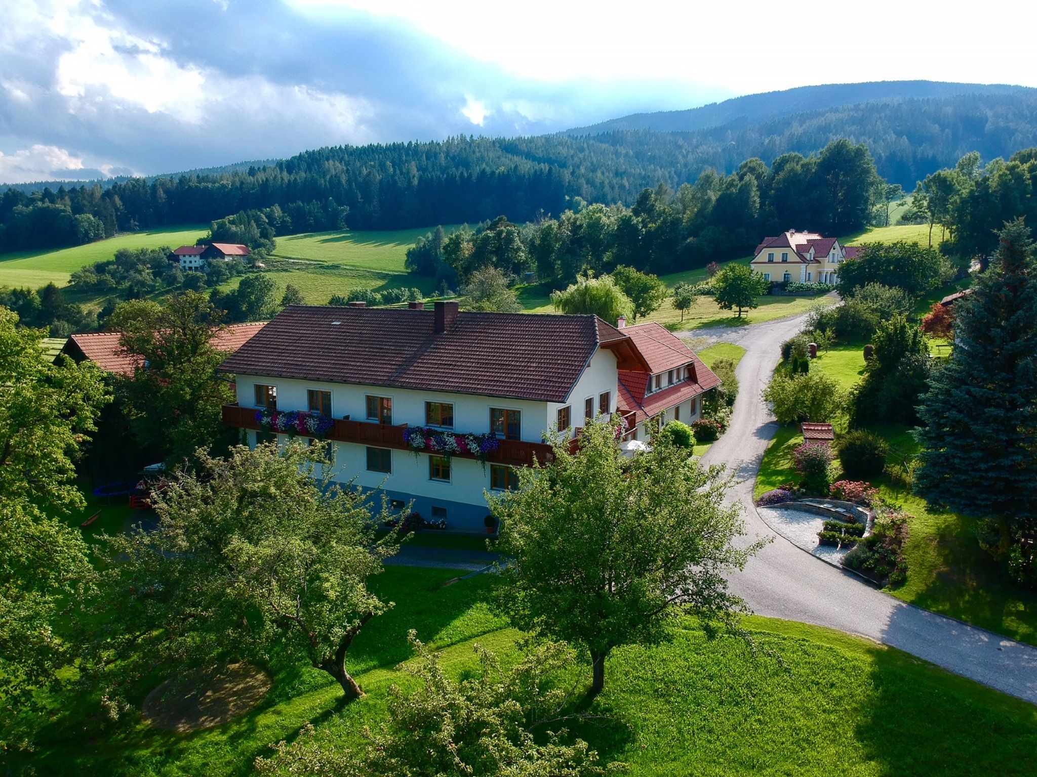 Vidiecka krajina s veľkým domom, obklopená stromami a lúkami, pod zamračenou oblohou.