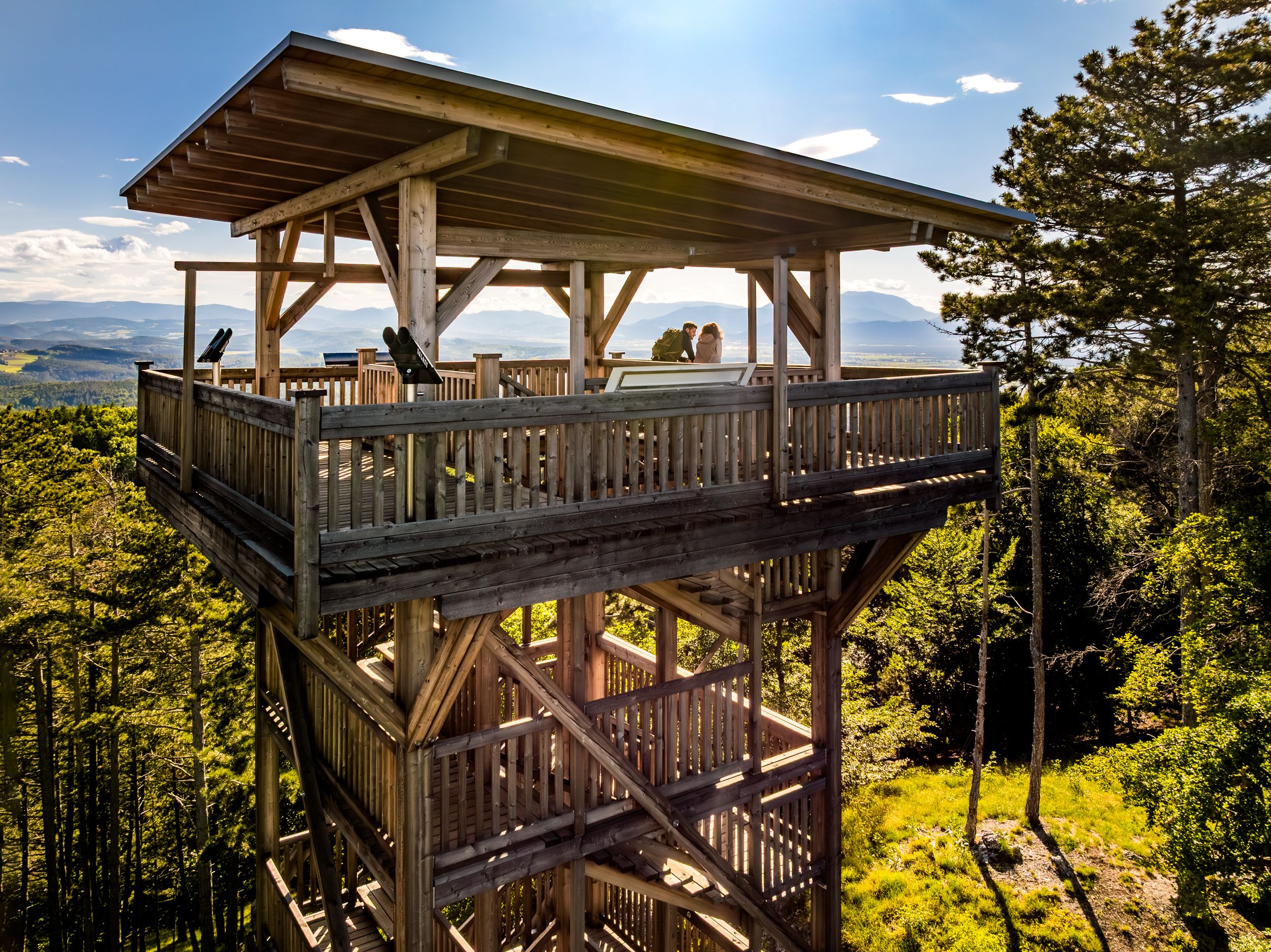 Záber z dronu zo strany vyhliadkovej veže, na ktorej stoja dvaja ľudia. Zelený les a hory v pozadí