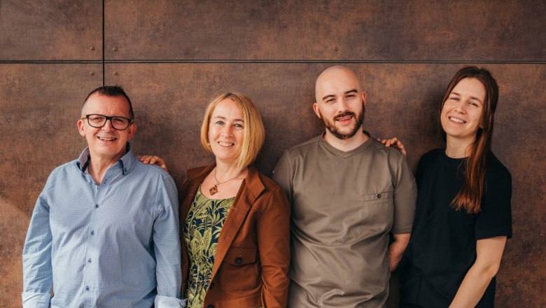 Štyria ľudia stoja s úsmevom vedľa seba pred hnedou stenou.