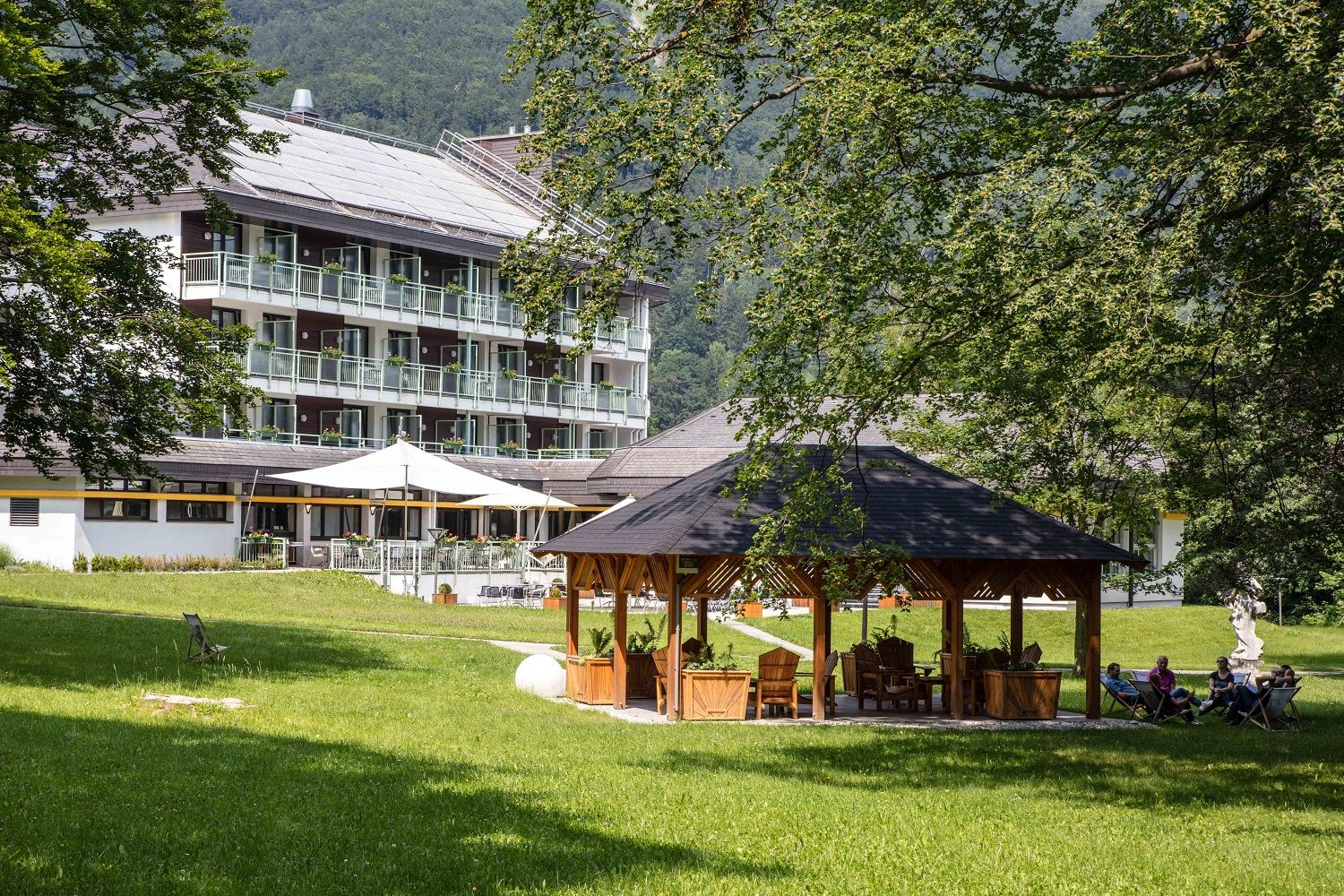 Hotelová budova s balkónmi v pozadí, pavilón a ľudia na stoličkách v popredí, obklopené stromami a trávnikmi.