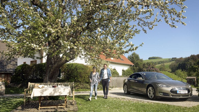 Pár kráča vedľa zaparkovaného elektromobilu pod rozkvitnutým stromom vo vidieckej záhrade.