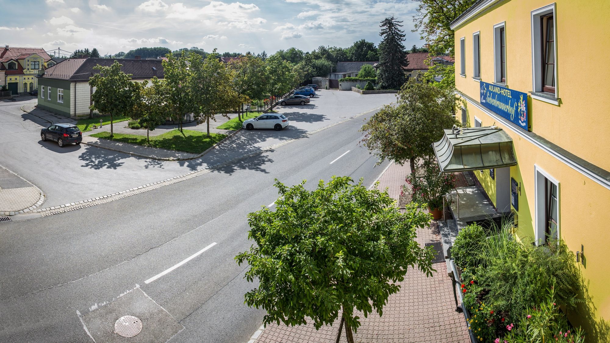 Scéna ulice so žltou budovou a parkoviskom, stromy a autá.