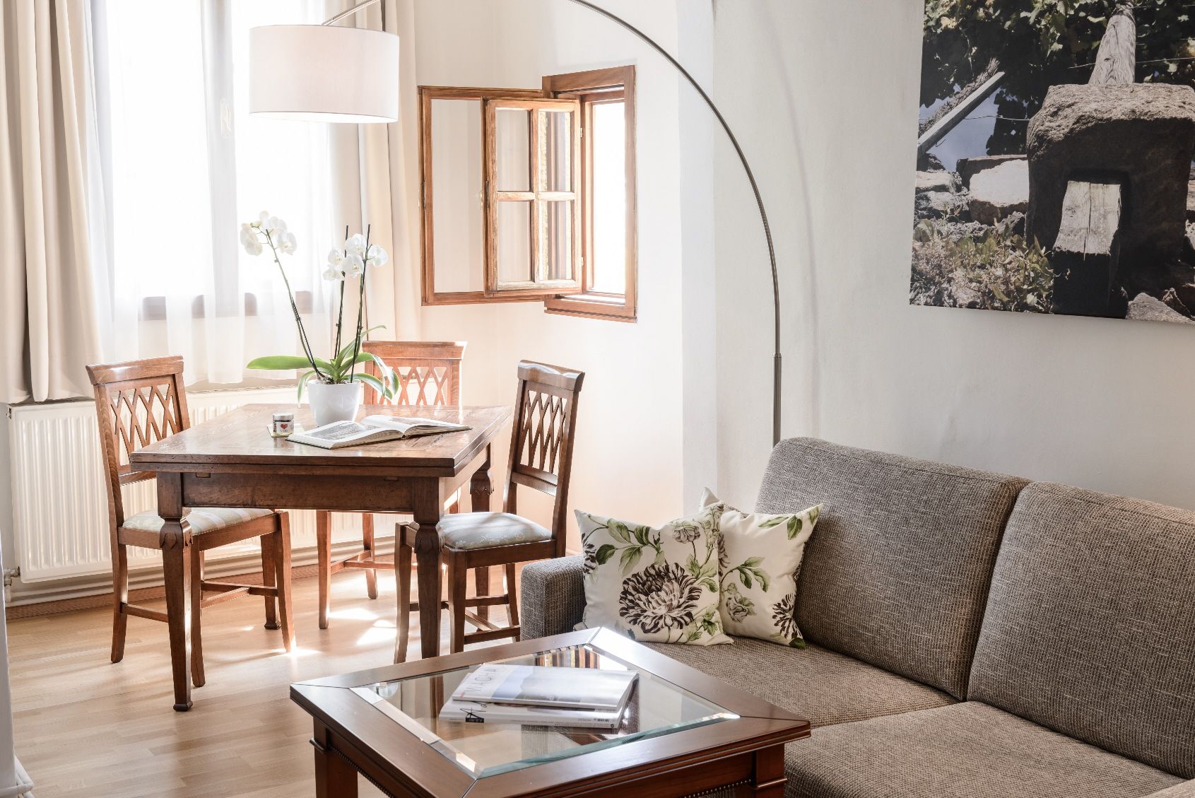 Útulná obývacia izba so sivou pohovkou, dreveným stolom a stoličkami, svetlým oknom a rastlinami.