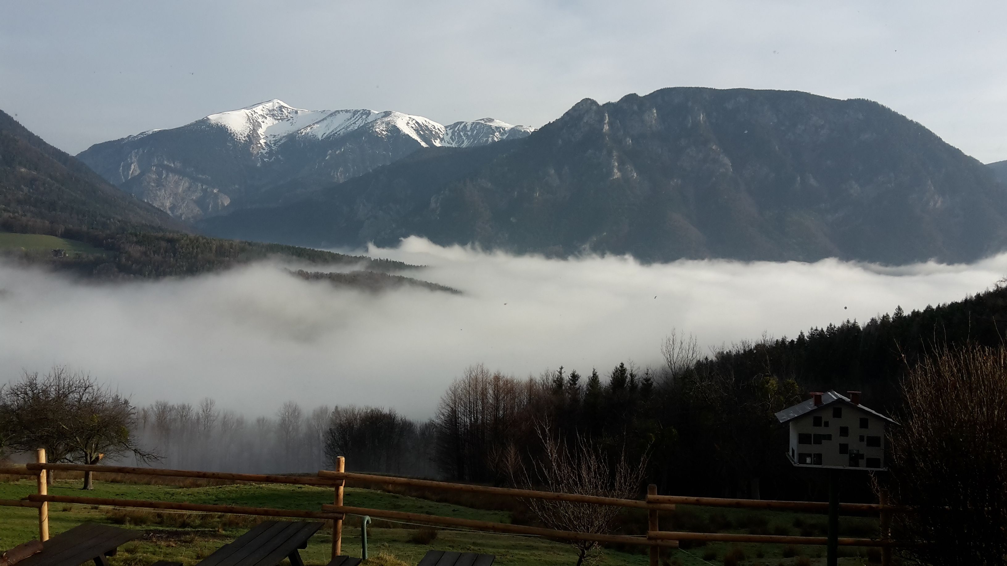 Pohľad na zasnežené hory nad údolím s hmlou a dreveným plotom v popredí.
