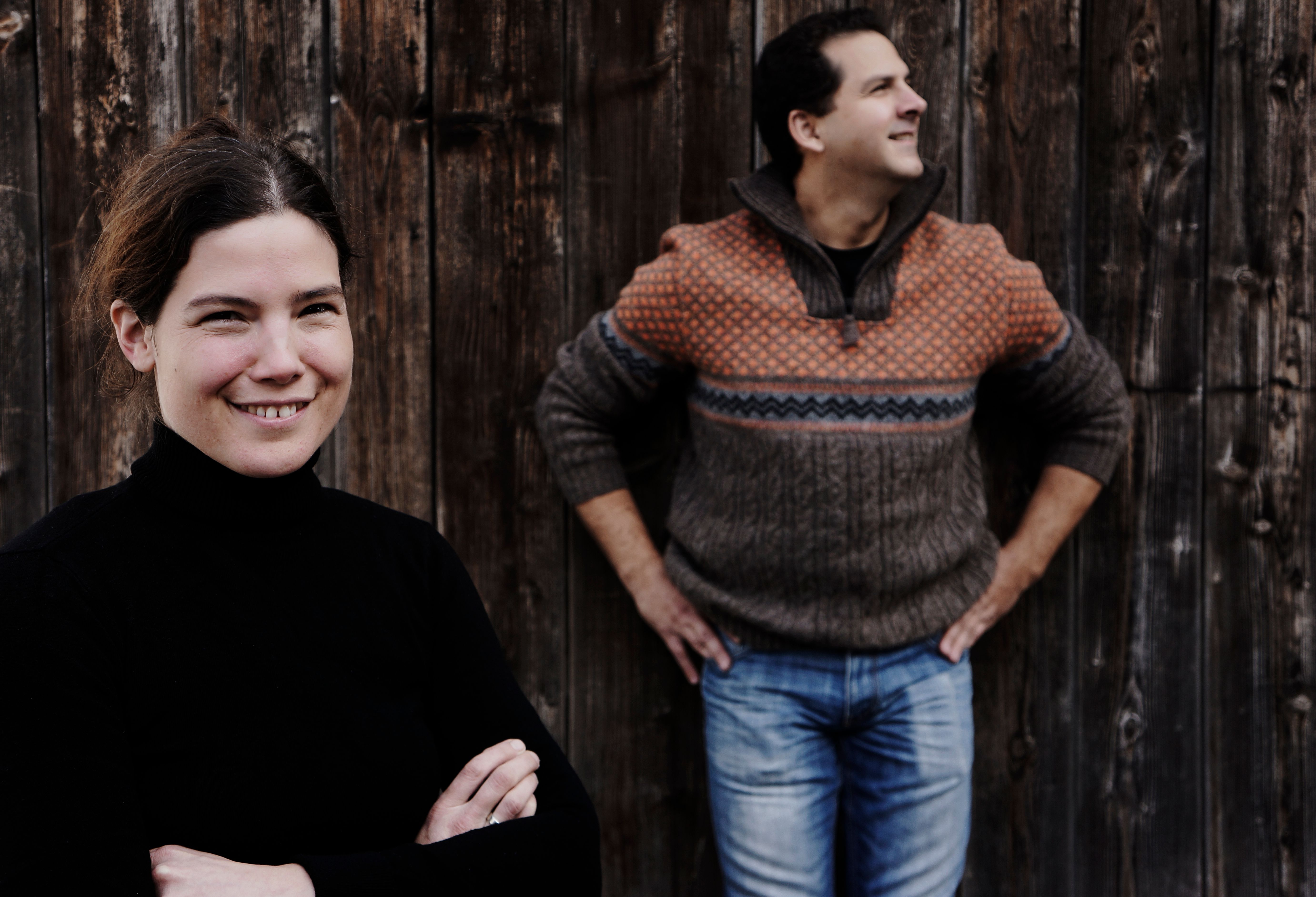 Dvaja ľudia pred drevenou stenou, žena v popredí sa usmieva, muž v pozadí sa pozerá do strany.
