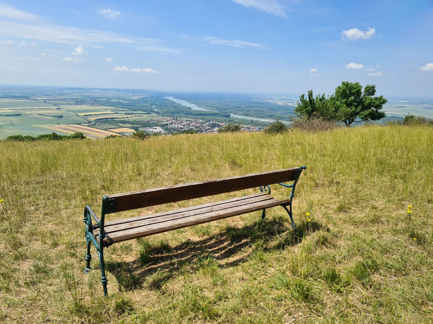 Drevená lavička na lúke s výhľadom na rozľahlú krajinu a rieku v diaľke.