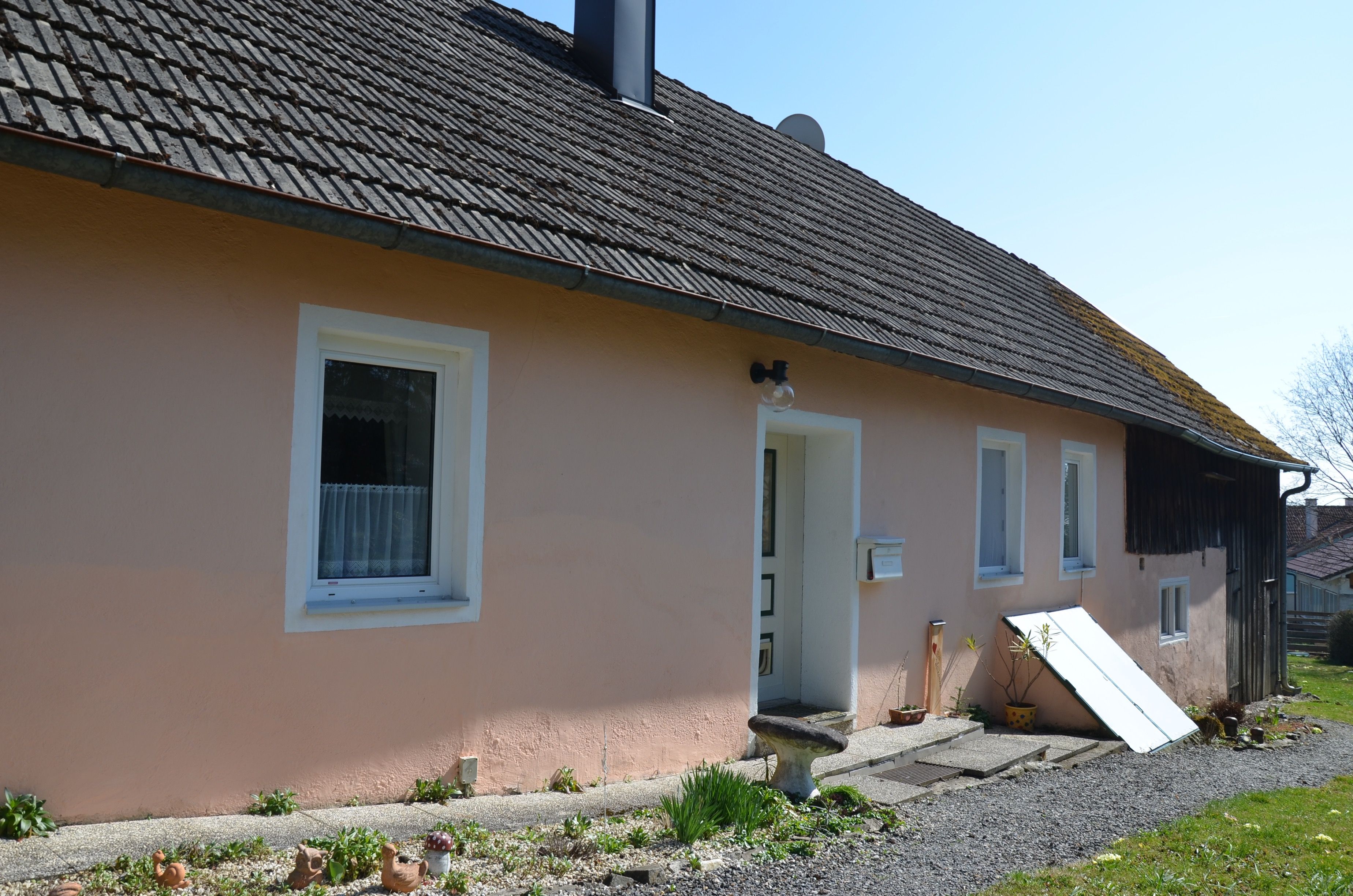 Jednoduchý jednopodlažný dom s ružovou fasádou a sivou strechou. Pred domom možno vidieť rastliny a dekorácie.