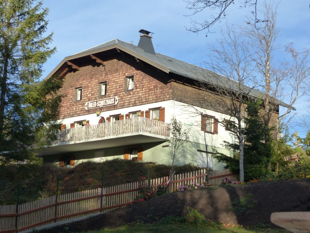 Tradičná budova s dreveným obložením a balkónom, obklopená stromami a dreveným plotom. Nápis: "Döhler Shelter".