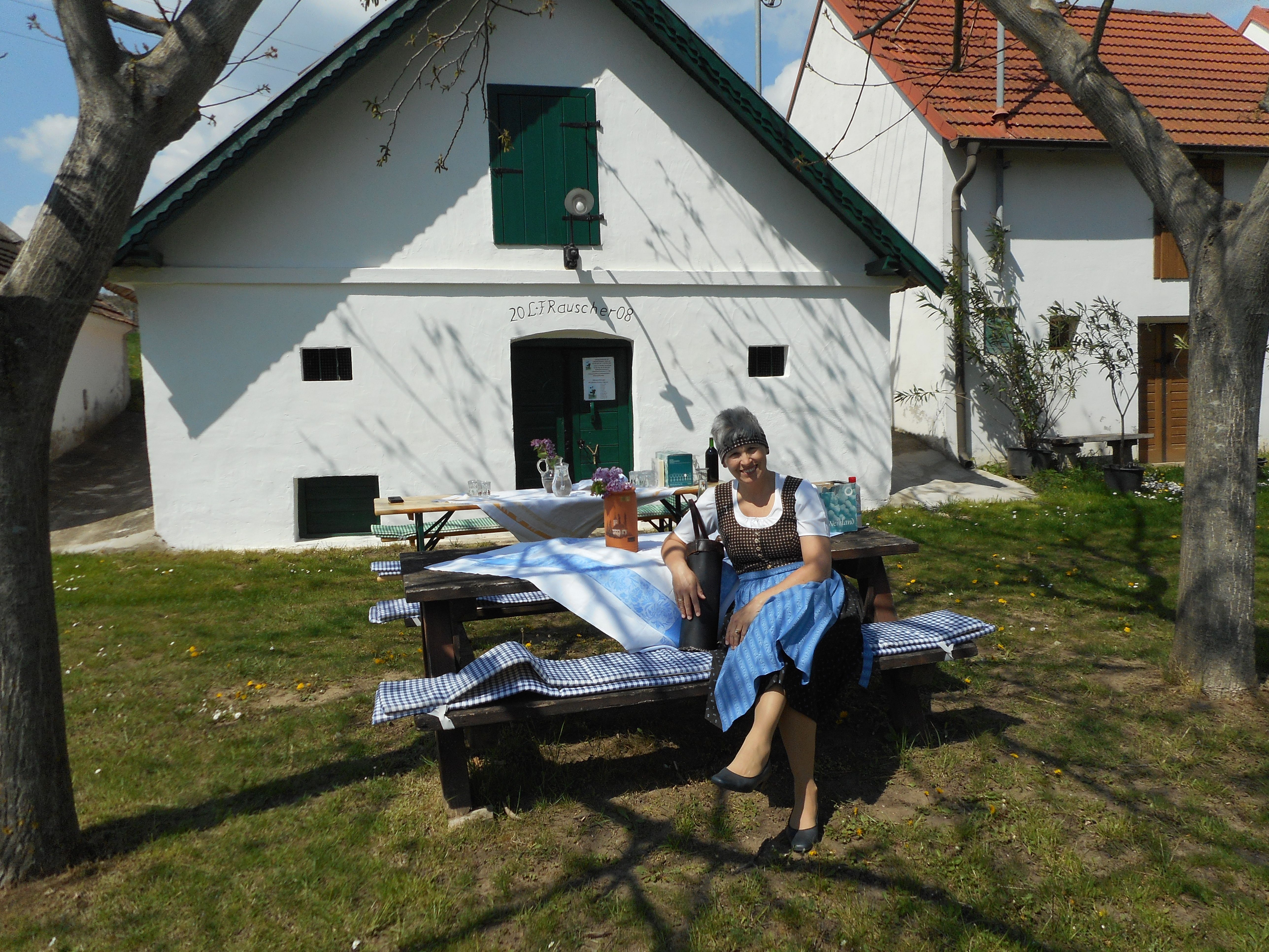 Žena v tradičnom odeve sedí na lavičke pred bielou budovou so zelenými okenicami.