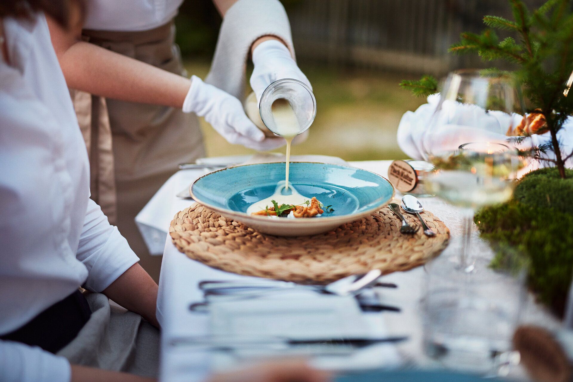Ein eleganter Tisch ist liebevoll gedeckt und lädt dazu ein, die kulinarischen Köstlichkeiten der Region zu genießen. Ein Kellner serviert mit Hingabe eine feine Suppe, während die umgebende Natur eine friedliche Atmosphäre schafft, die zum Verweilen einlädt.