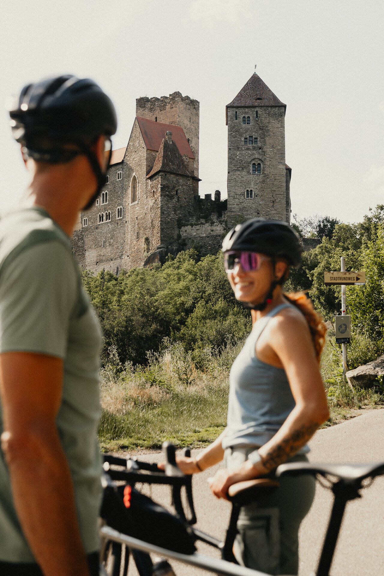Im Vordergrund etwas unscharf eine Radfahrerin, die einen Radfahrer an, hinter ihr die Burg Hardegg scharfgestellt