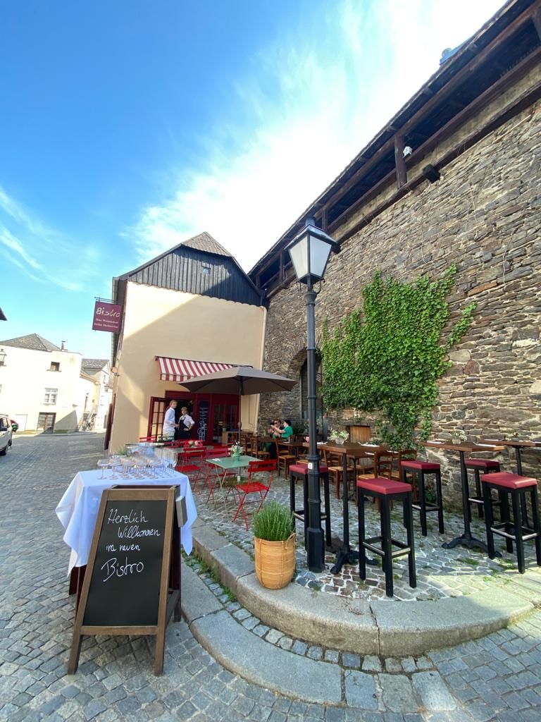 Útulné bistro s červenými stoličkami a stolmi vonku, obklopené starými kamennými múrmi a rastlinami.