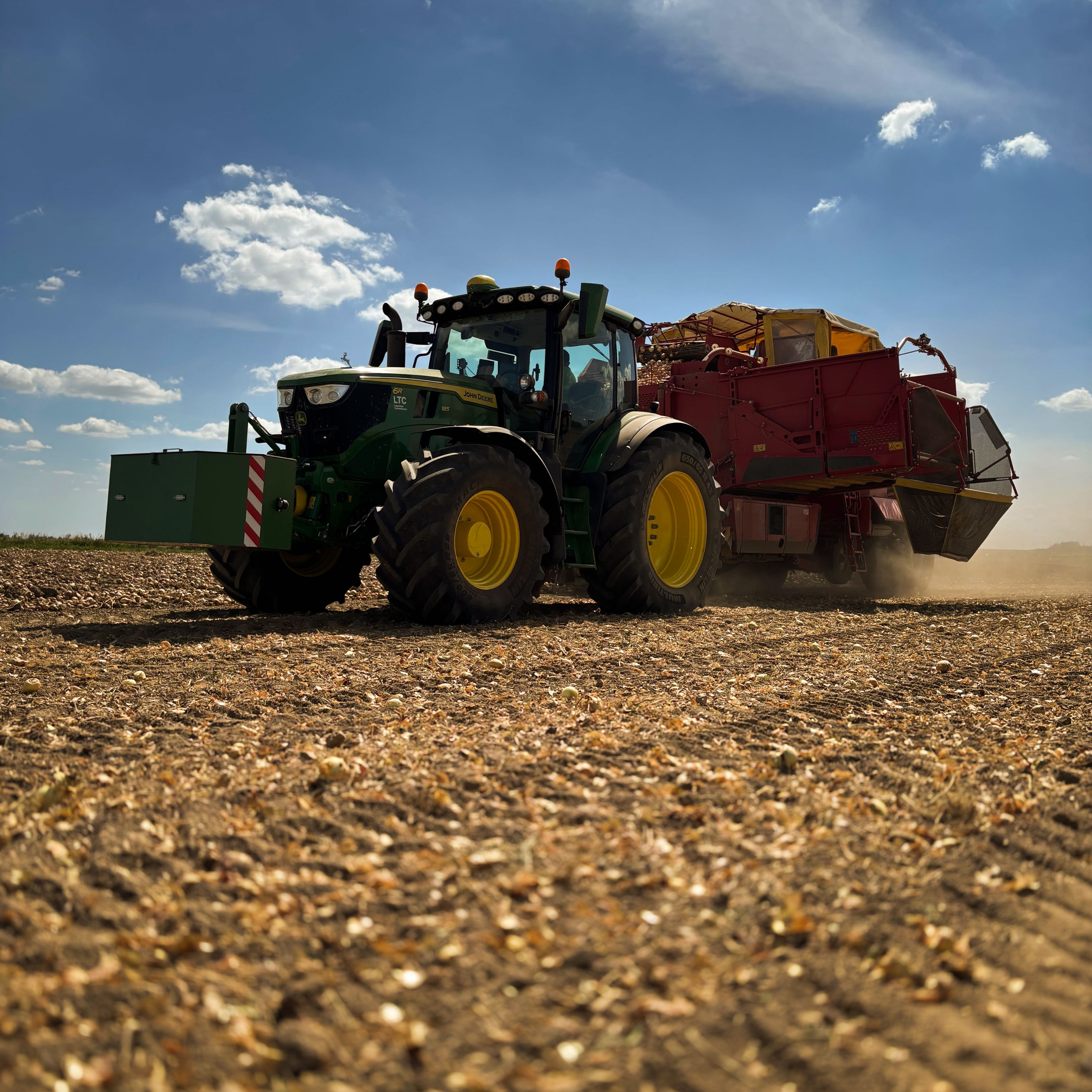 Traktor s prívesom prechádza cez obrobené pole pod modrou oblohou s niekoľkými mrakmi.