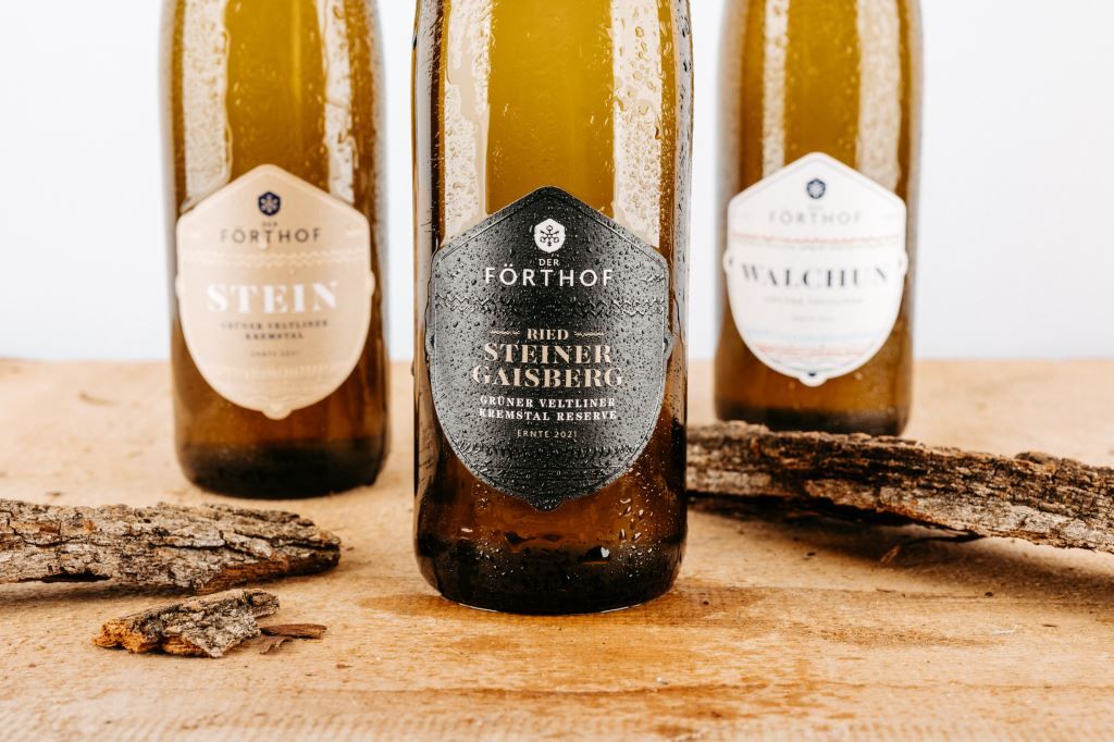 Tri fľaše vína Förthof na drevenom stole s kúskami kôry.