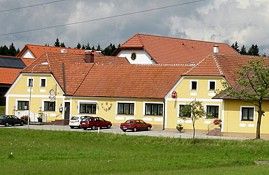 Žltá budova s červenými strechami a niekoľkými autami pred ňou, obklopená zelenými lúkami a stromami.