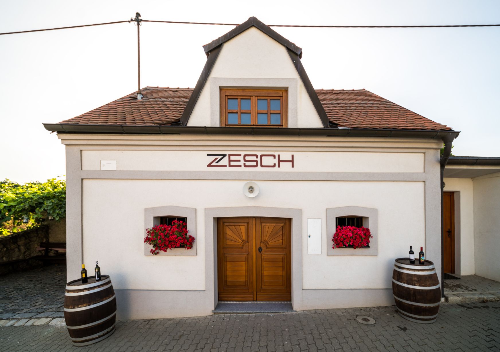 Malá budova s nápisom "Zesch", dva sudy na víno a červené kvety.