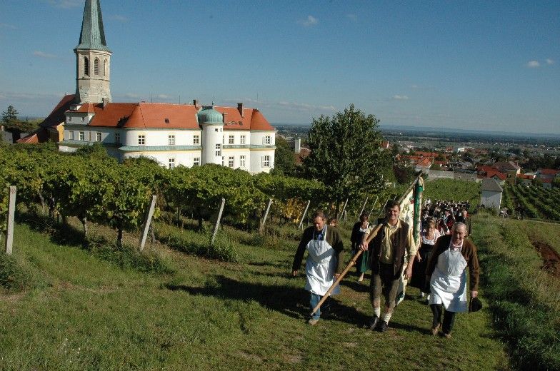 Ľudia v tradičných krojoch prechádzajú vinohradmi v Gumpoldskirchene, v pozadí kostol a budovy.