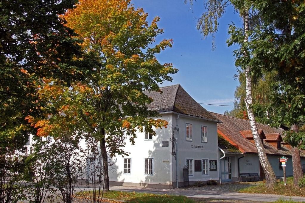 Biela budova s nápisom "Heimatmuseum", obklopená stromami s jesenným lístím.