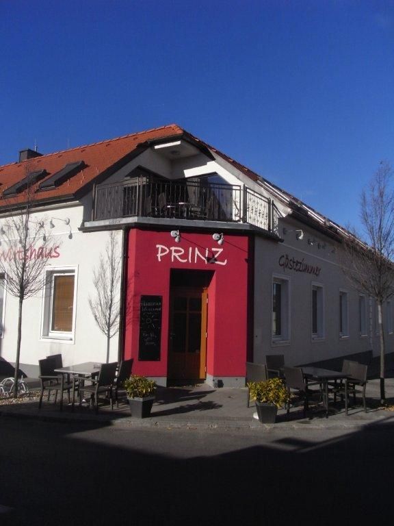 Pohľad zvonku na budovu s označením "Prinz", záhrada pre hostí so stolmi a stoličkami, modrá obloha.