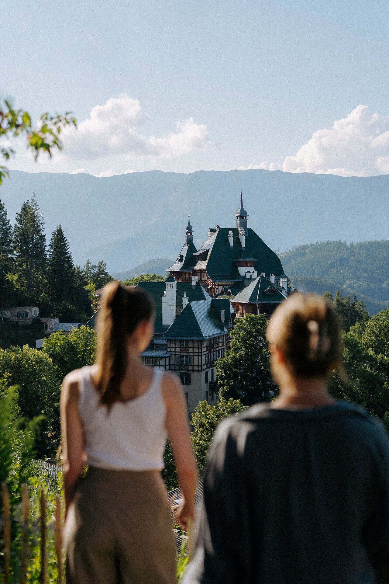 Dvaja ľudia otočení chrbtom ku kamere sa pozerajú na malebnú horskú krajinu. V popredí je vidieť hotel Südbahnhotel, ktorý je zasadený do sviežej zelene. V pozadí sa pod jasnou modrou oblohou dvíhajú hory.