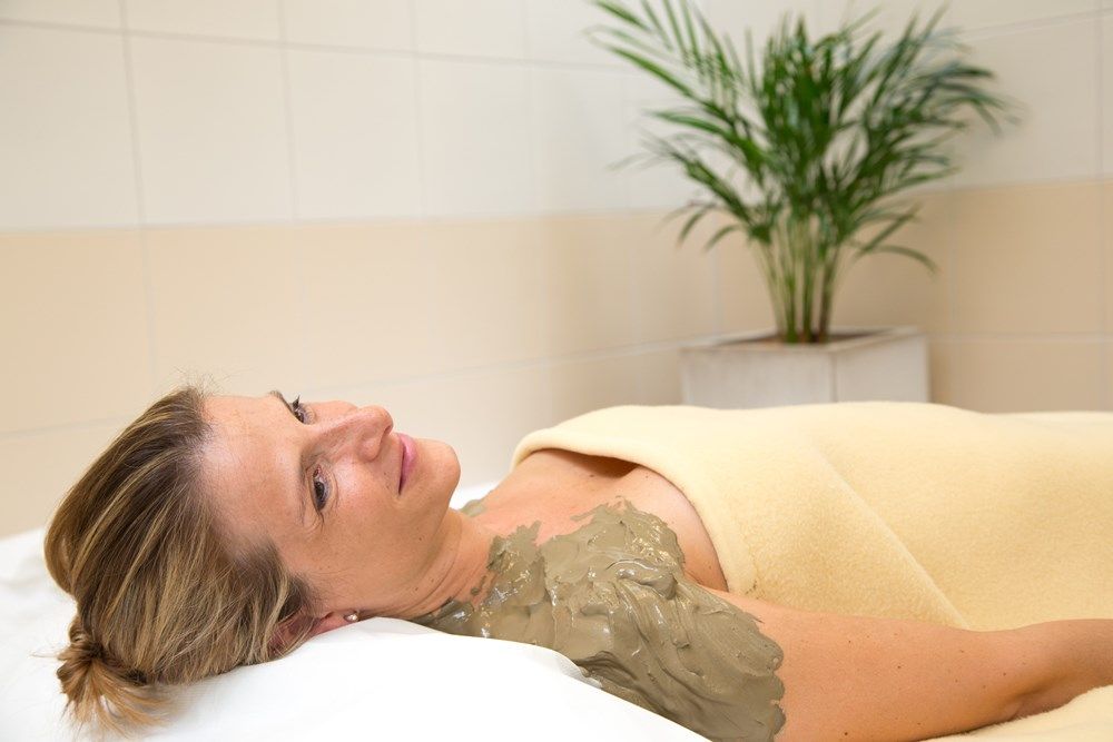 Žena relaxuje so zábalom zo sírneho bahna na ramene, zabalená v deke, vo wellness zóne s rastlinou v pozadí.