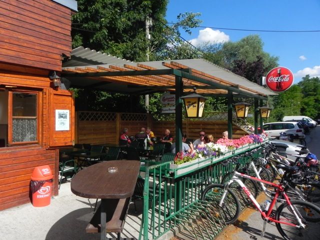 Vonkajší priestor snack baru s drevenými stenami, stolmi a bicyklami. Ľudia sedia pod zastrešeným priestorom. Viditeľný nápis Coca-Cola.