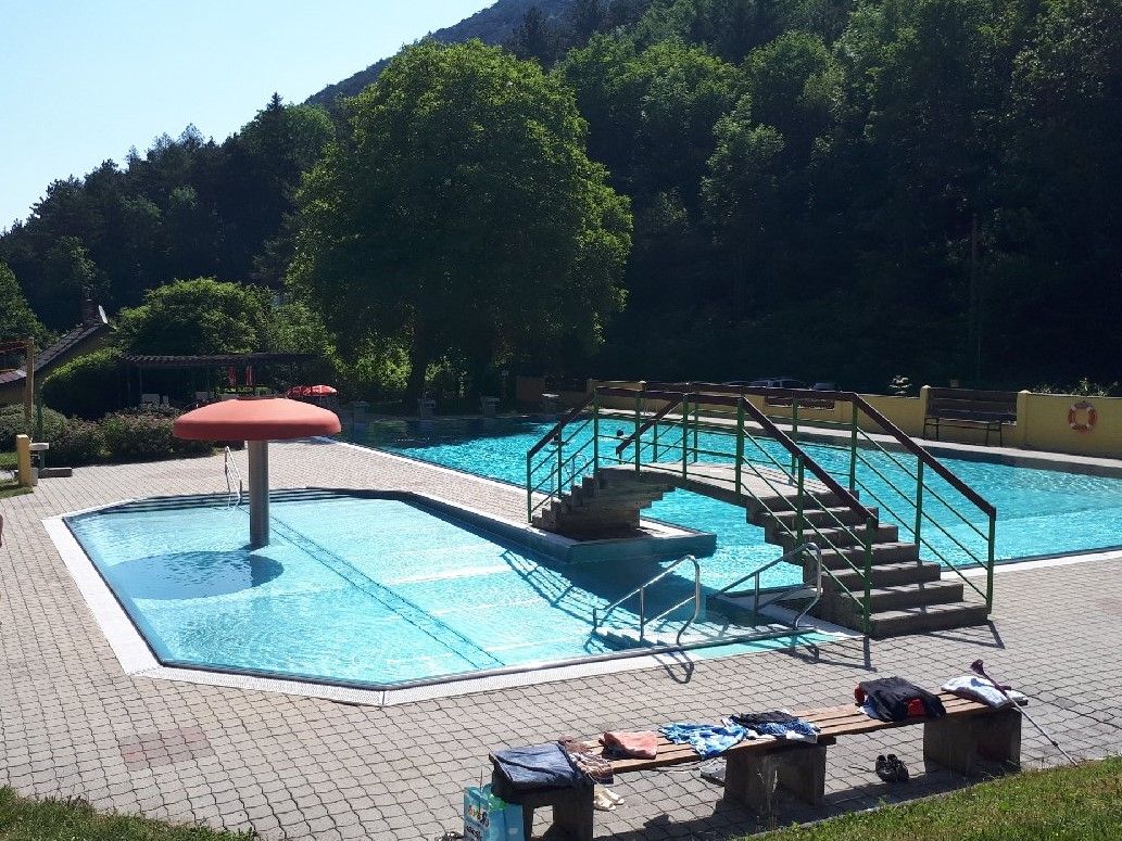 Vonkajší bazén s malým bazénom, mostíkom a hríbovým vodotryskom. Obklopený stromami a slnečnou oblohou.