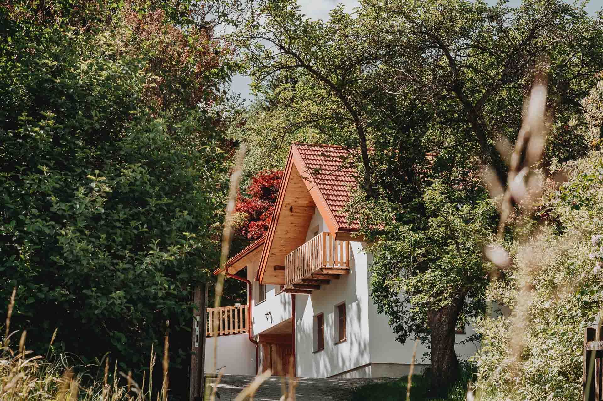Biely dom s červenou škridlovou strechou, obklopený stromami a rastlinami.